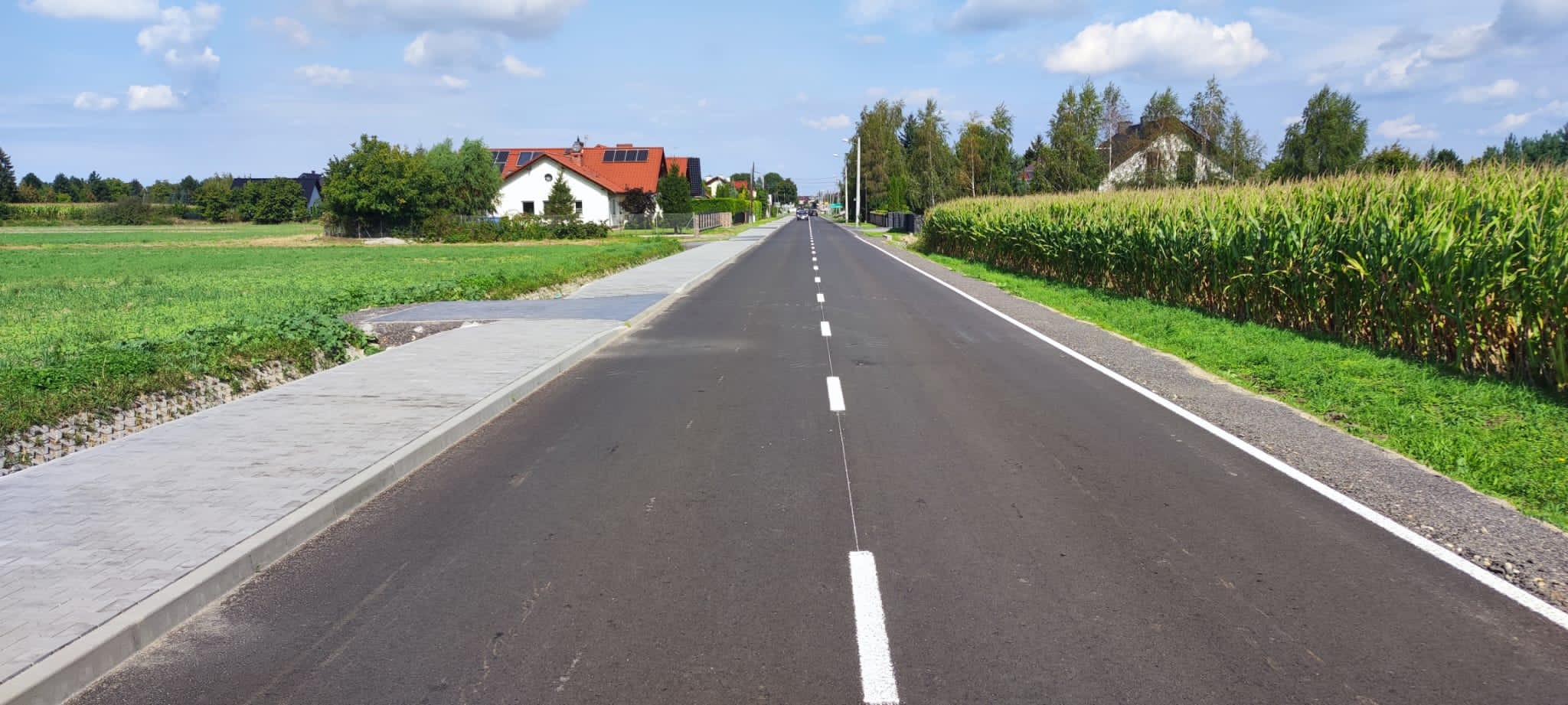 Asfaltowa droga z nowym oznakowaniem poziomym, po lewej stronie chodnik z kostki brukowej, w tle zabudowa jednorodzinna i pole kukurydzy.