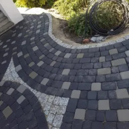 Ułożona ścieżka z ciemnej kostki brukowej z dekoracyjnymi wstawkami z jasnej kostki granitowej, obrzeże z drobnego kamienia, w tle zwinięty wąż ogrodowy i zieleń.