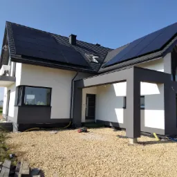Nowoczesny dom jednorodzinny z ciemnoszarą dachówką i panelami fotowoltaicznymi, widoczne obróbki blacharskie i okno dachowe, teren wokół domu jeszcze w trakcie prac wykończeniowych.