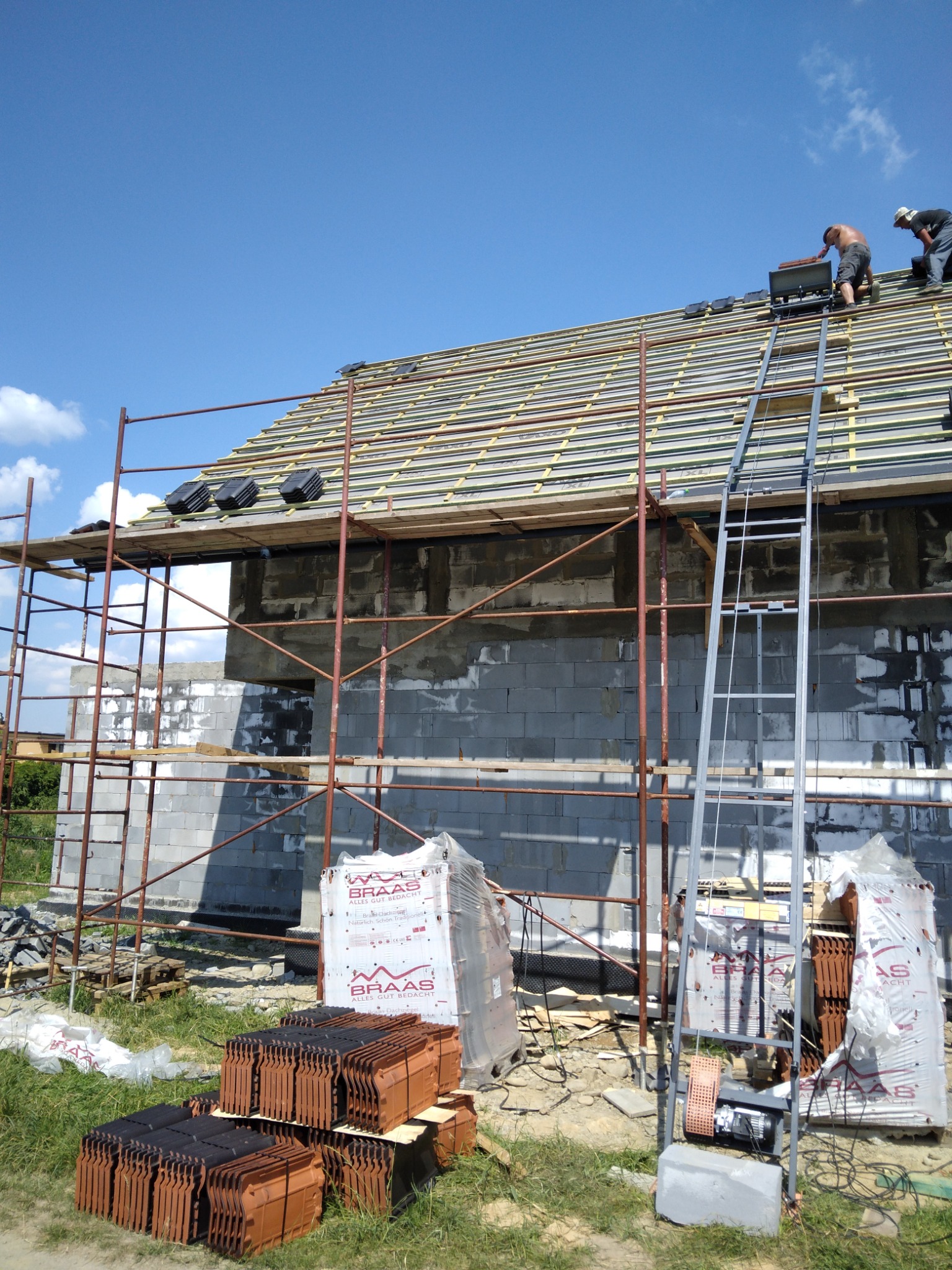 Montaż brązowej dachówki Braas na nowym dachu budynku w trakcie budowy, widoczne rusztowania i podnośnik do dachówki, dwóch dekarzy na dachu, błękitne niebo.