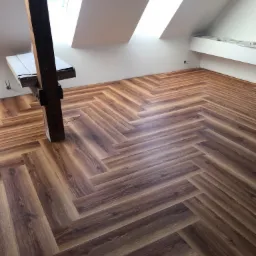 Ukończona podłoga w jodełkę z paneli drewnopodobnych w jasnym poddaszu z drewnianą belką konstrukcyjną. Widoczne skosy i okna dachowe.