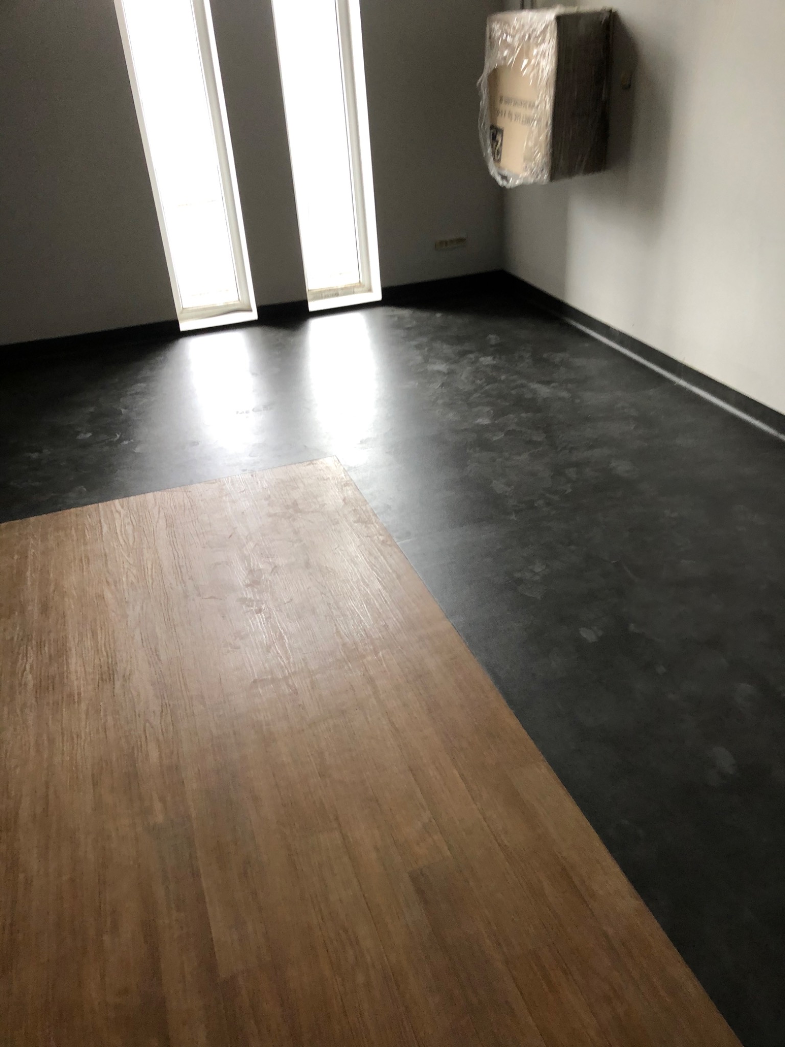 Fragment ułożonej podłogi z paneli drewnopodobnych, widoczne także fragmenty szarej podłogi i ściany z oknami oraz karton przy suficie.