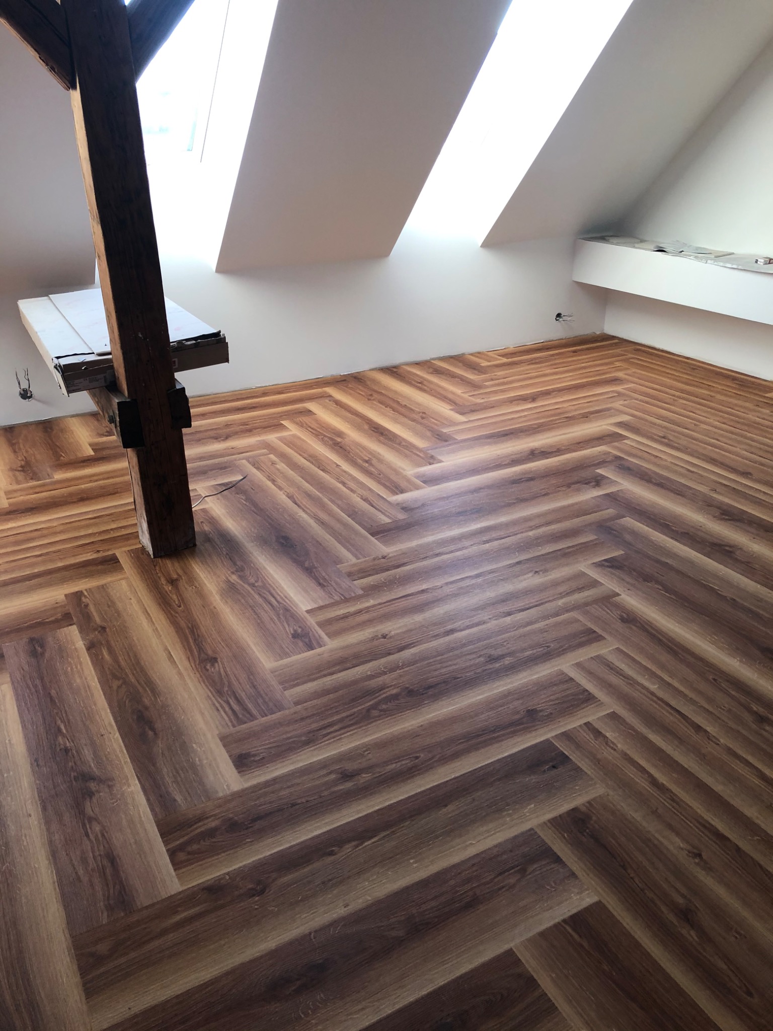 Ukończona podłoga w jodełkę z paneli drewnopodobnych w jasnym poddaszu z drewnianą belką konstrukcyjną. Widoczne skosy i okna dachowe.