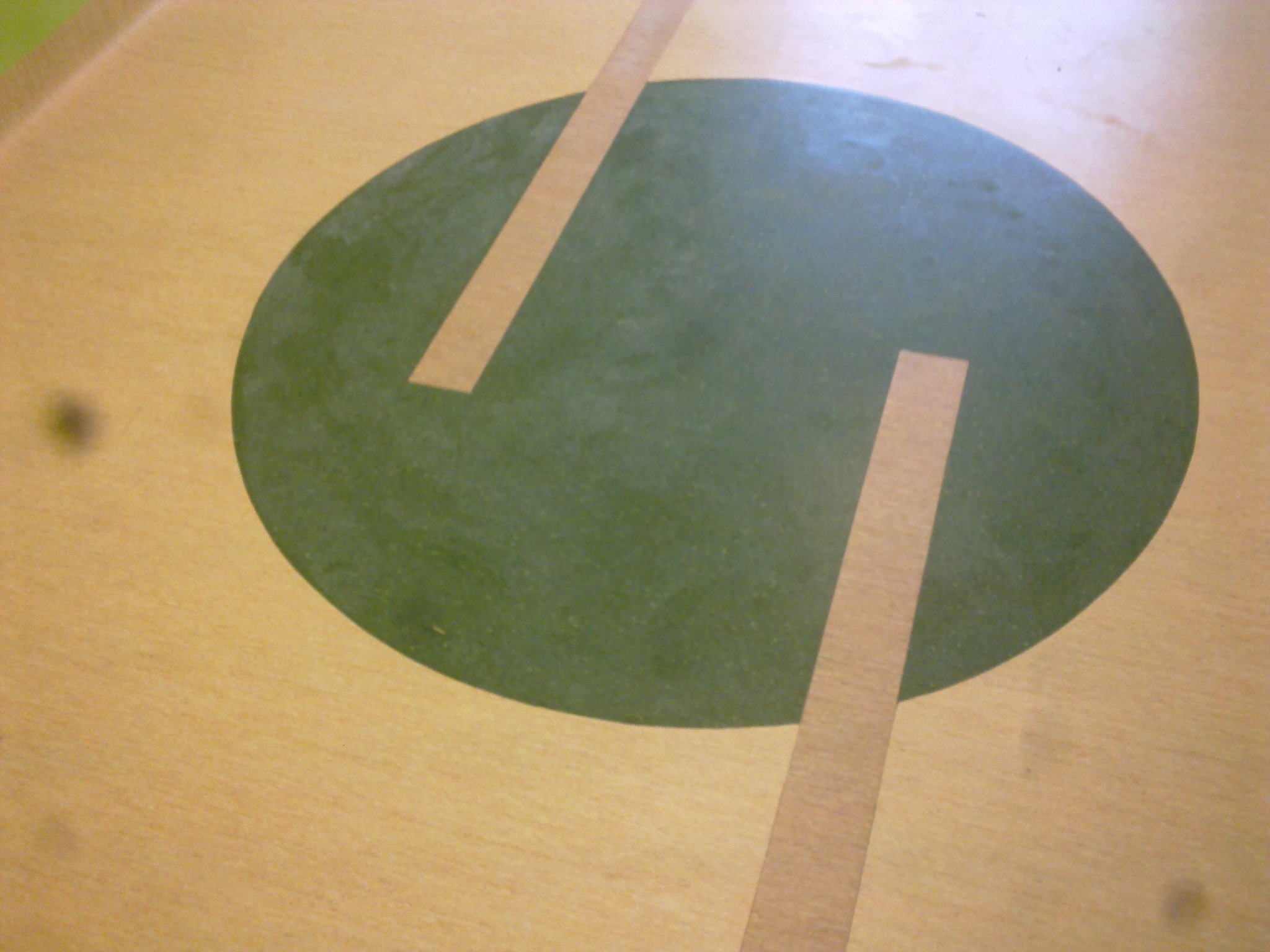 Zbliżenie na podłogę z jasnego drewna z wkomponowanym ciemnozielonym owalem, przeciętym dwiema cienkimi, jasnymi paskami.