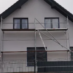 Dom jednorodzinny w trakcie prac elewacyjnych, widoczne rusztowanie, okna zabezpieczone folią, świeżo nałożony tynk w kolorze jasnoszarym, czarny dach i orynnowanie.