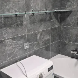 Szara łazienka z marmurowymi płytkami, szklaną półką na chromowanych wspornikach, białą wanną i pralką Electrolux z podłączonym przewodem zasilającym.