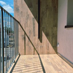 Remontuje Świdwin. Pl - Nowoczesny balkon z podłogą i ścianą wyłożoną jasnymi płytkami imitującymi drewno, z ciemną metalową balustradą i ostrymi, ukośnymi cieniami. W tle błękitne niebo.