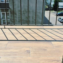 Remontuje Świdwin. Pl - Balkon z jasnymi płytkami drewnopodobnymi i cieniami balustrad, z widokiem na szarą fasadę budynku z oknami i roletami, w słonecznym świetle.