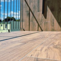 Remontuje Świdwin. Pl - Słoneczny balkon z podłogą i ścianą wyłożoną jasnymi płytkami imitującymi drewno, z czarną balustradą i ostrymi cieniami słońca.