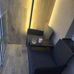 Remontuje Świdwin. Pl - Nowoczesny balkon z ciemnymi meblami z technorattanu, podłogą i pionową ścianą z płytek drewnopodobnych. Zintegrowane, ciepłe oświetlenie LED podkreśla fakturę i wieczorny nastrój.