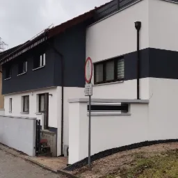 Dwukondygnacyjny dom z elewacją w kolorach bieli i grafitu, widoczny znak drogowy na pierwszym planie, pochmurne niebo w tle.