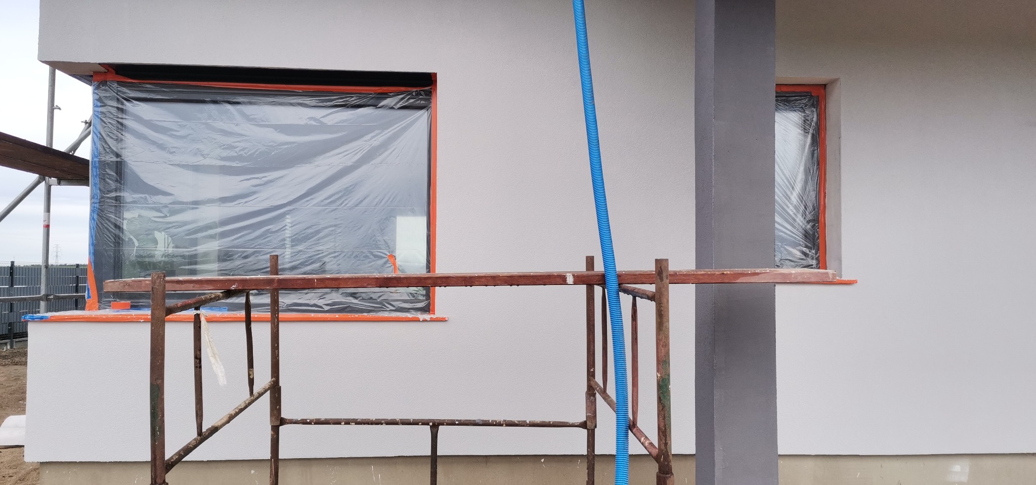 Dom w trakcie prac elewacyjnych: okna zabezpieczone folią, widoczne rusztowanie i świeżo otynkowana, jasnoszara ściana.