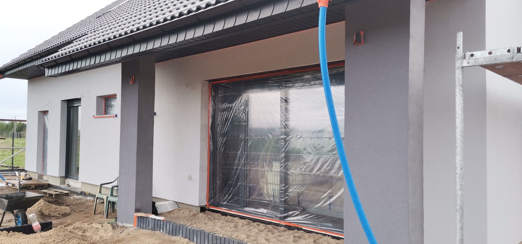 Fragment elewacji domu w trakcie prac wykończeniowych, z oknami zabezpieczonymi folią, widoczny fragment rusztowania i niebieski przewód wentylacyjny.