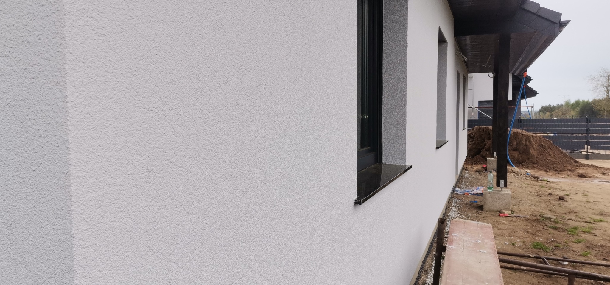 Jasnoszara elewacja domu jednorodzinnego z oknami w czarnych ramach i czarnymi parapetami z kamienia, widok z boku na tle ziemi i drzew.