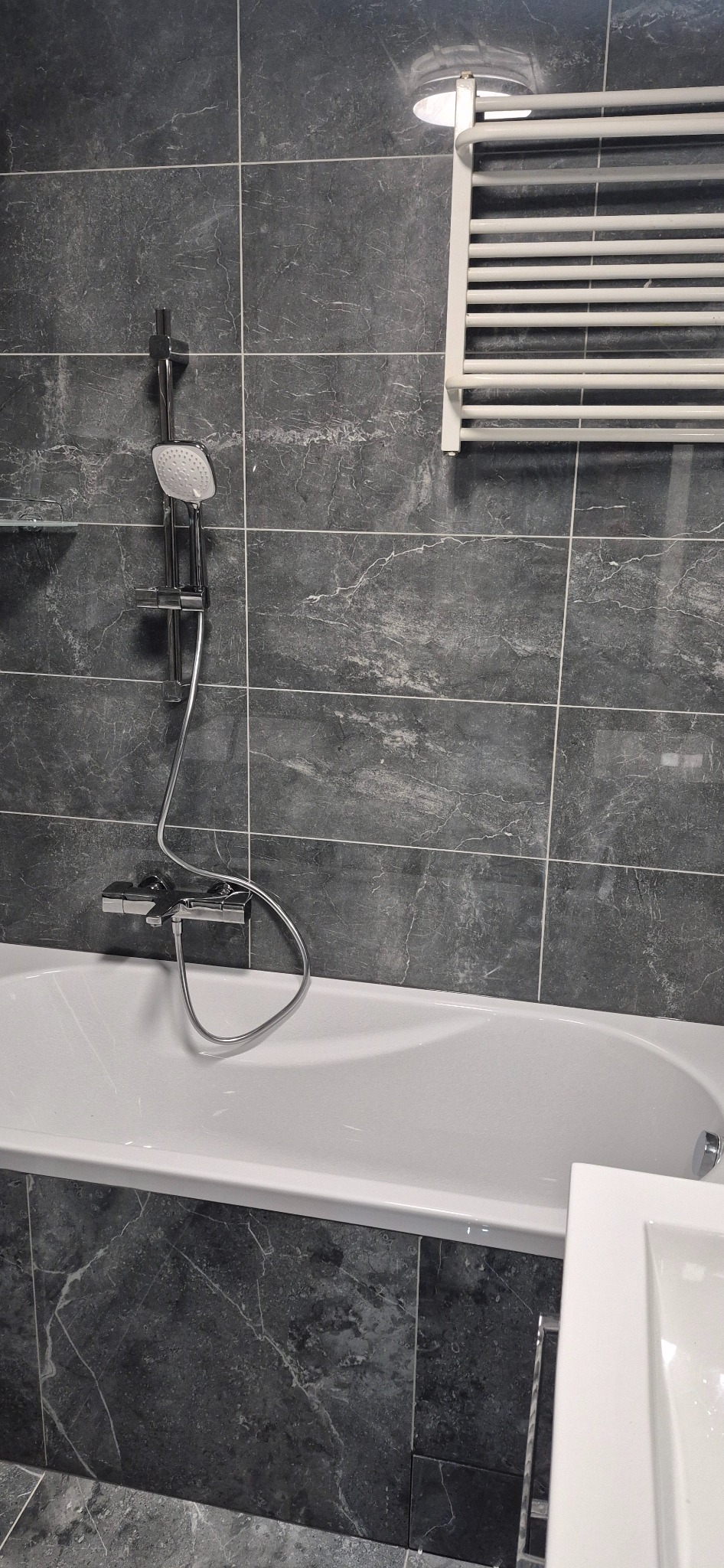 Nowoczesna łazienka z wanną, szarą marmurkową glazurą i białym grzejnikiem drabinkowym, oświetlona górnym światłem.