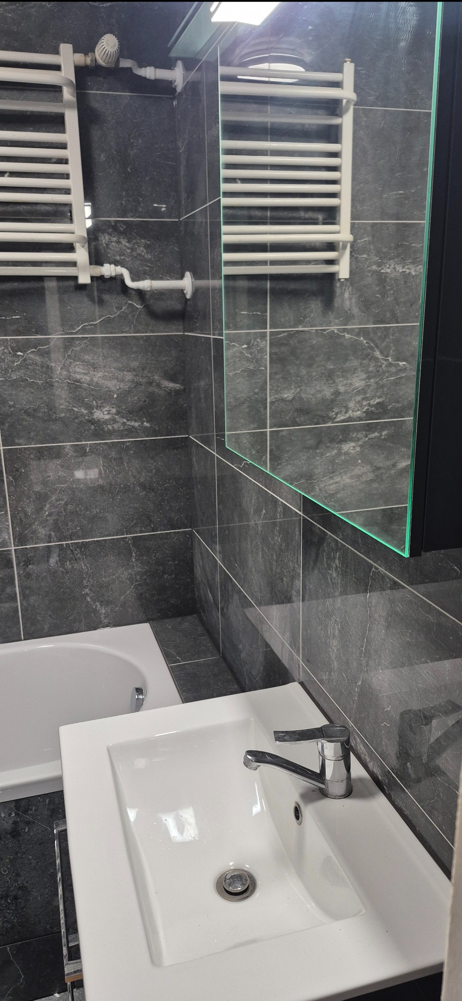 Nowoczesna łazienka z szarymi płytkami imitującymi marmur, białą umywalką z chromowaną baterią i grzejnikiem drabinkowym, odbicie w lustrze.