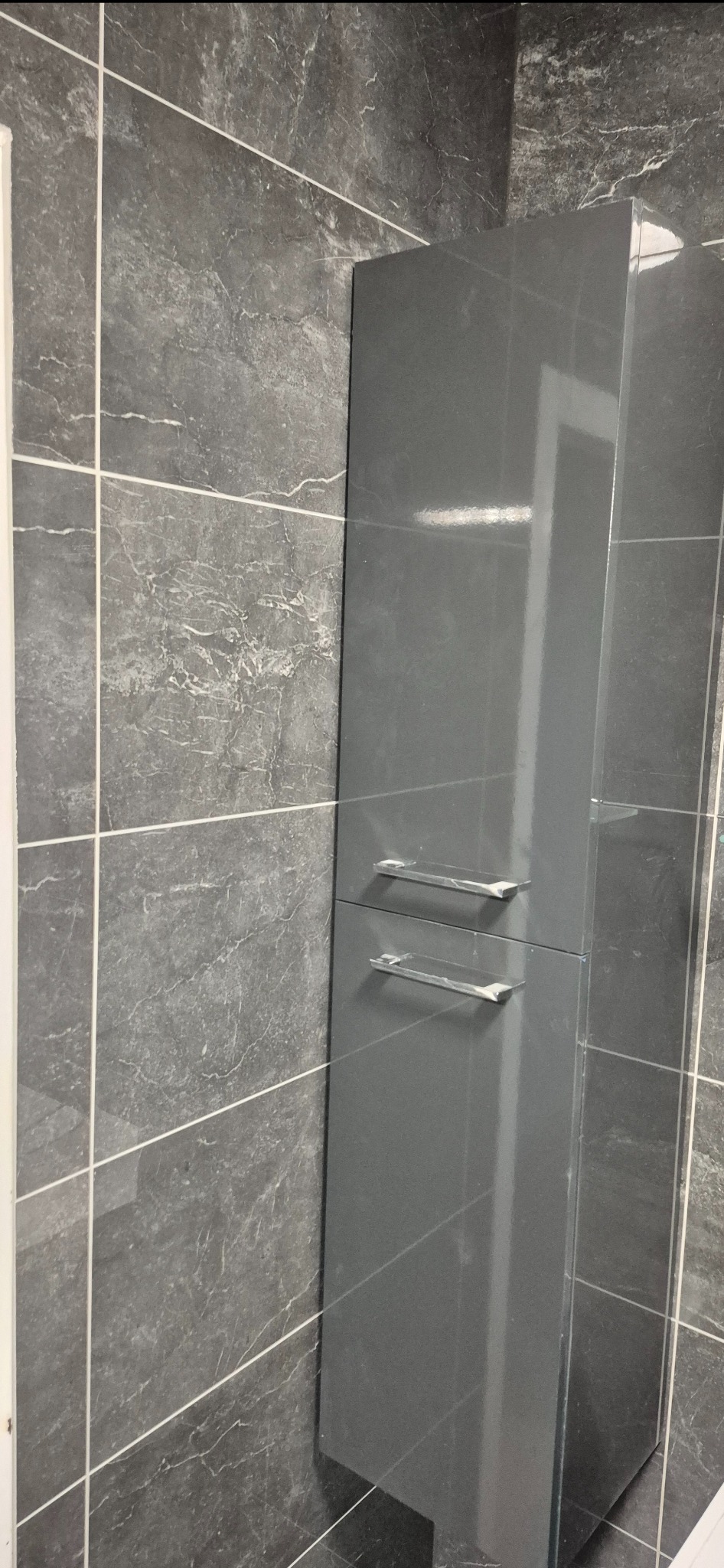 Szara, lakierowana szafka łazienkowa z dwoma szufladami i chromowanymi uchwytami na tle ściany wyłożonej płytkami imitującymi szary marmur z białymi fugami.