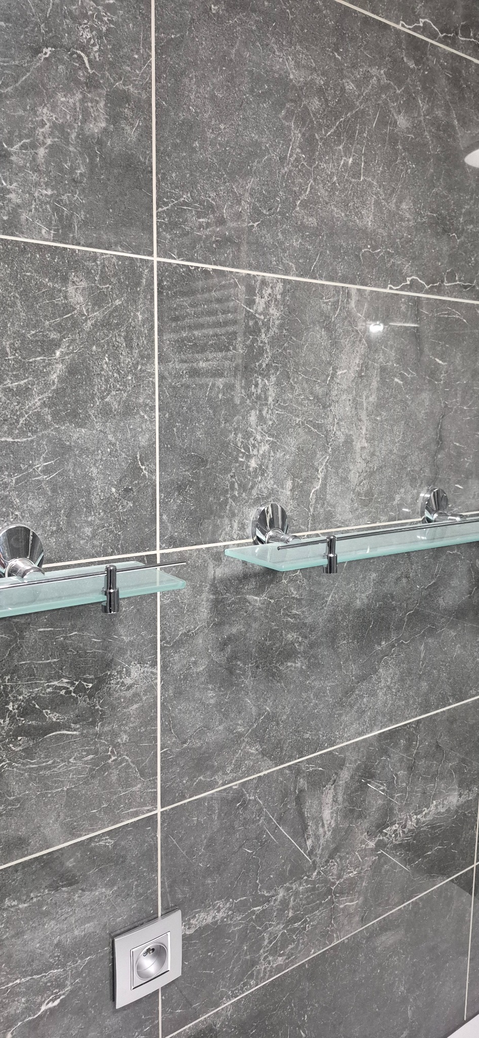 Szara ściana z płytek imitujących marmur, z zamontowanymi szklanymi półkami na chromowanych wspornikach i gniazdkiem elektrycznym.