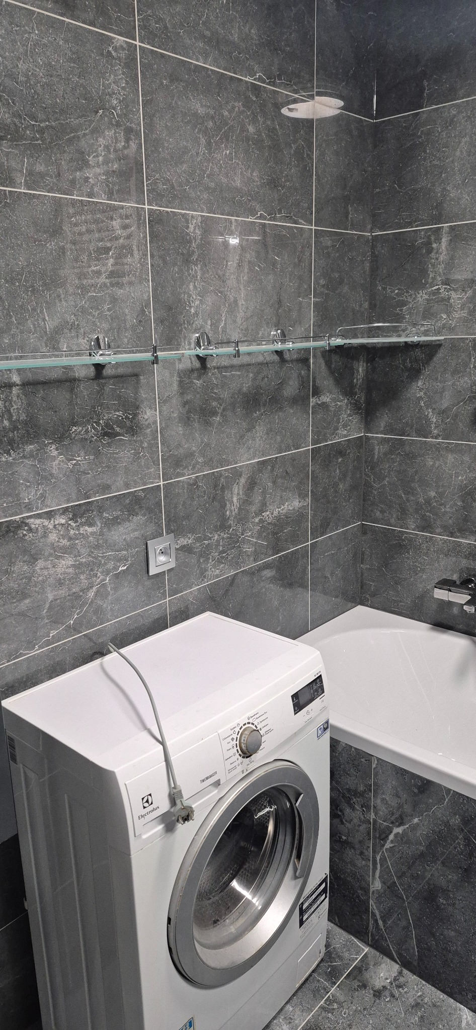 Szara łazienka z marmurowymi płytkami, szklaną półką na chromowanych wspornikach, białą wanną i pralką Electrolux z podłączonym przewodem zasilającym.