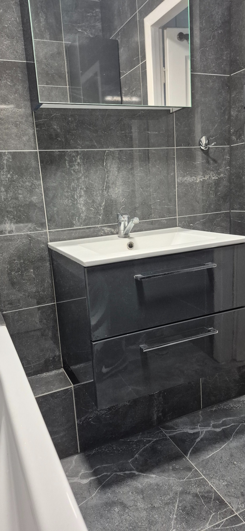 Czarna łazienka z umywalką w szafce z dwoma szufladami, szare marmurowe płytki na ścianach i podłodze, oraz lustro z szafką wiszące nad umywalką.