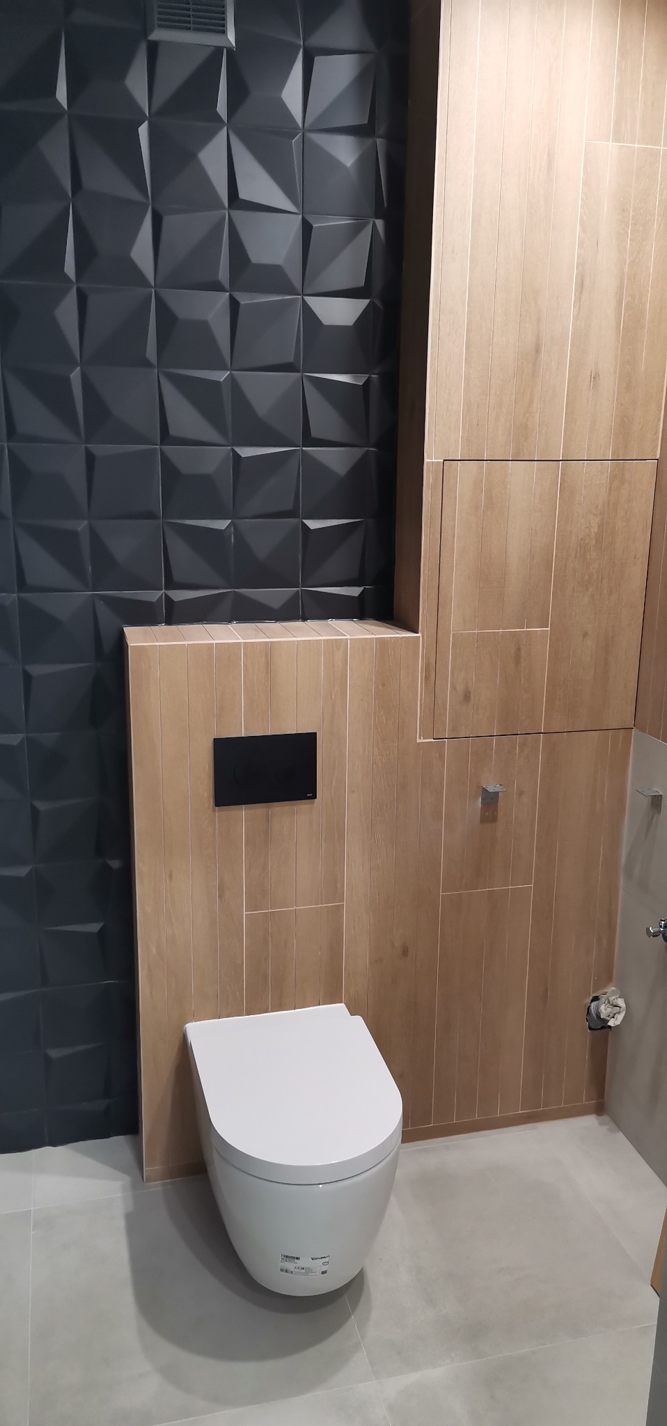 Nowoczesna toaleta podwieszana z czarnym panelem sterującym na ścianie wyłożonej płytkami imitującymi drewno, obok ściana pokryta trójwymiarowymi panelami w odcieniach grafitu, szara podłoga.