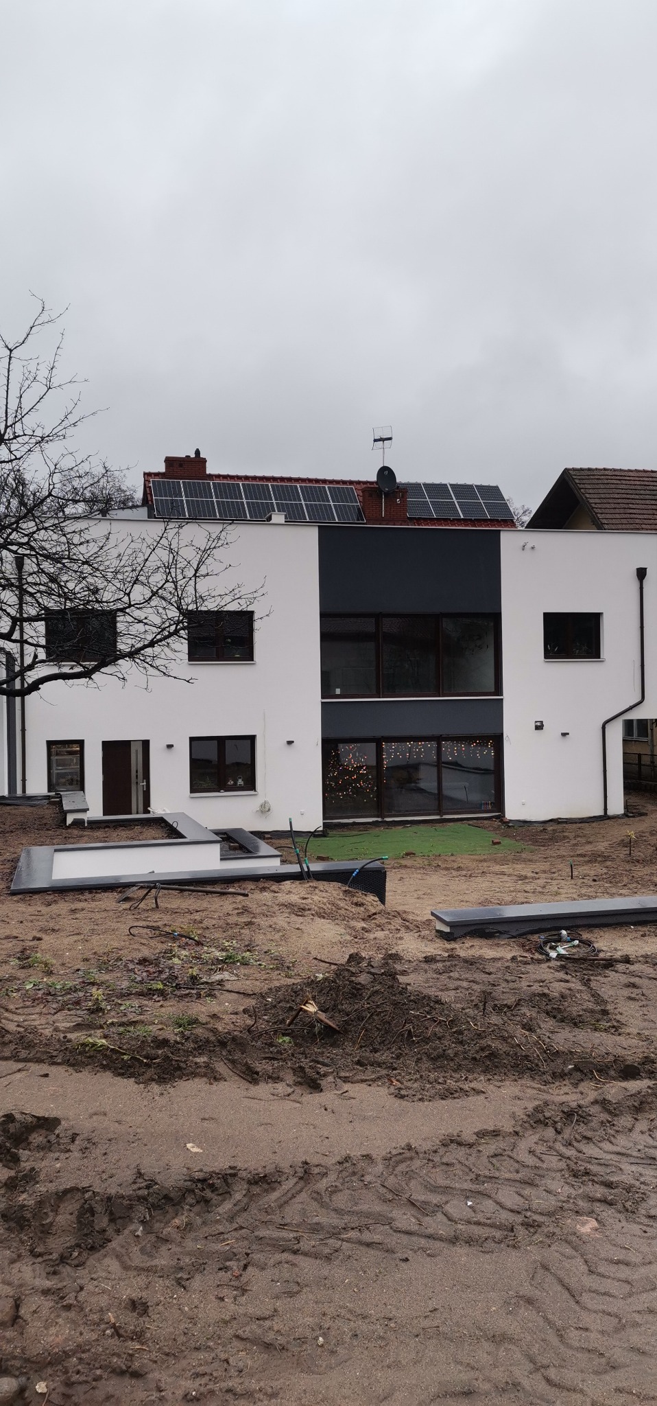 Dwukondygnacyjny dom z panelami słonecznymi na dachu, w trakcie prac wykończeniowych ogrodu, widoczne elementy obrzeży trawnikowych i ziemia z odciśniętymi śladami opon.