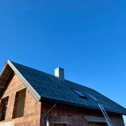 Niedokończony dom z cegły z zielonym dachem w trakcie budowy, widoczne rynny, okna dachowe, komin i drabina oparta o ścianę, na drugim planie błękitne niebo.