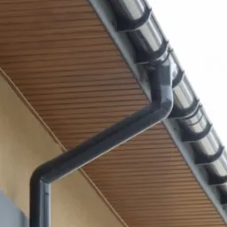 Szara, metalowa rynna spustowa, widoczna na tle brązowej podbitki dachowej i fragmentu elewacji budynku.