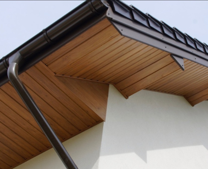 Fragment dachu z drewnianą podbitką i brązową rynną spustową, widok z dołu na jasnym tle ściany budynku.