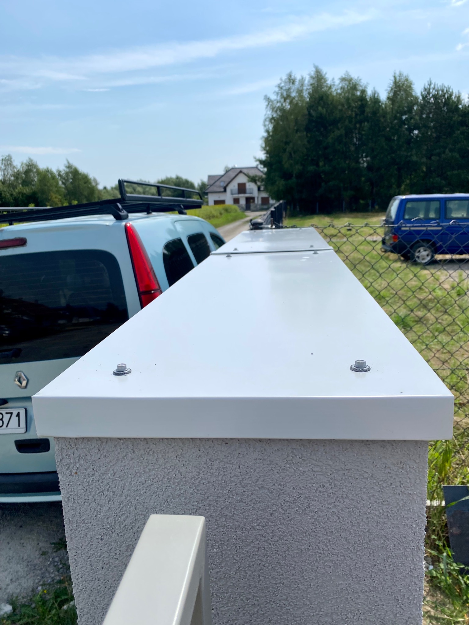 Szara, otynkowana podmurówka ogrodzenia w Medyni Głogowskiej z zamontowaną, jasną, metalową obróbką blacharską. W tle widoczne samochody i dom.