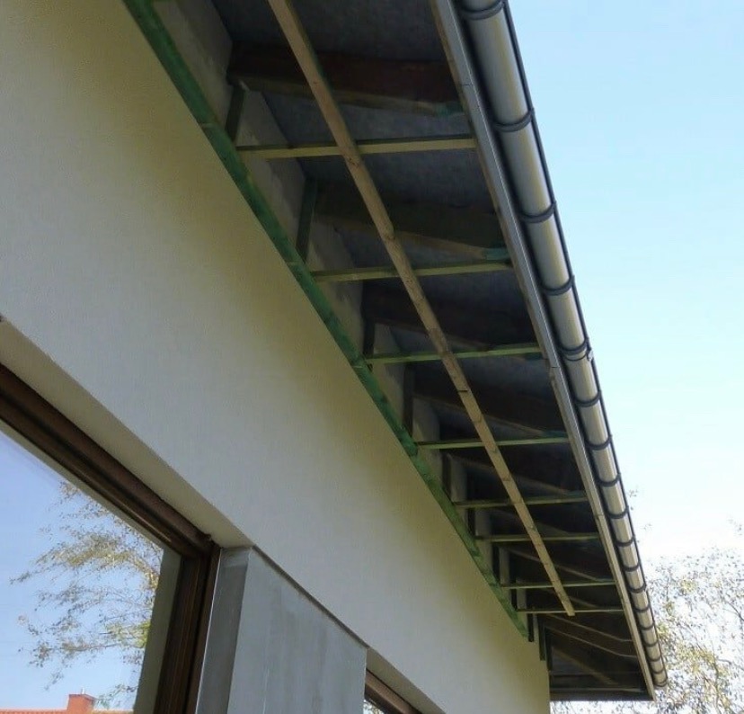 Wystający okap dachu z widoczną konstrukcją drewnianą, izolacją i zamontowaną szarą rynną plastikową na tle błękitnego nieba.