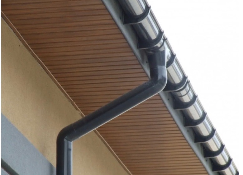 Szara, metalowa rynna spustowa, widoczna na tle brązowej podbitki dachowej i fragmentu elewacji budynku.