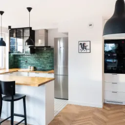 Nowoczesna kuchnia z drewnianym blatem, zielonymi płytkami na ścianie, czarnymi lampami wiszącymi i podłogą ułożoną w jodełkę.