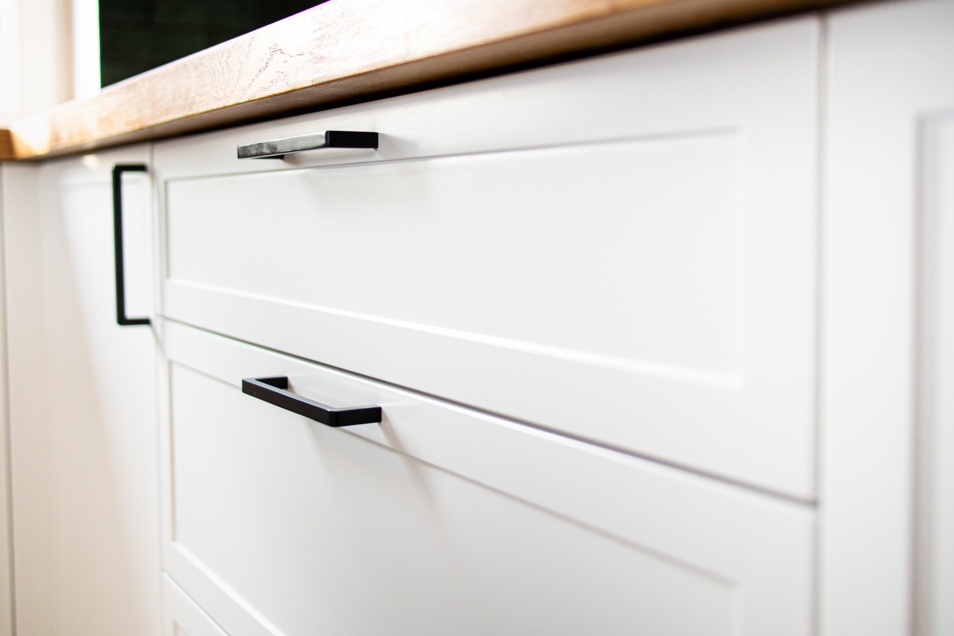 Białe, minimalistyczne fronty szafek z czarnymi, prostokątnymi uchwytami i drewnianym blatem, widok z bliska z lekkim kątem z dołu.