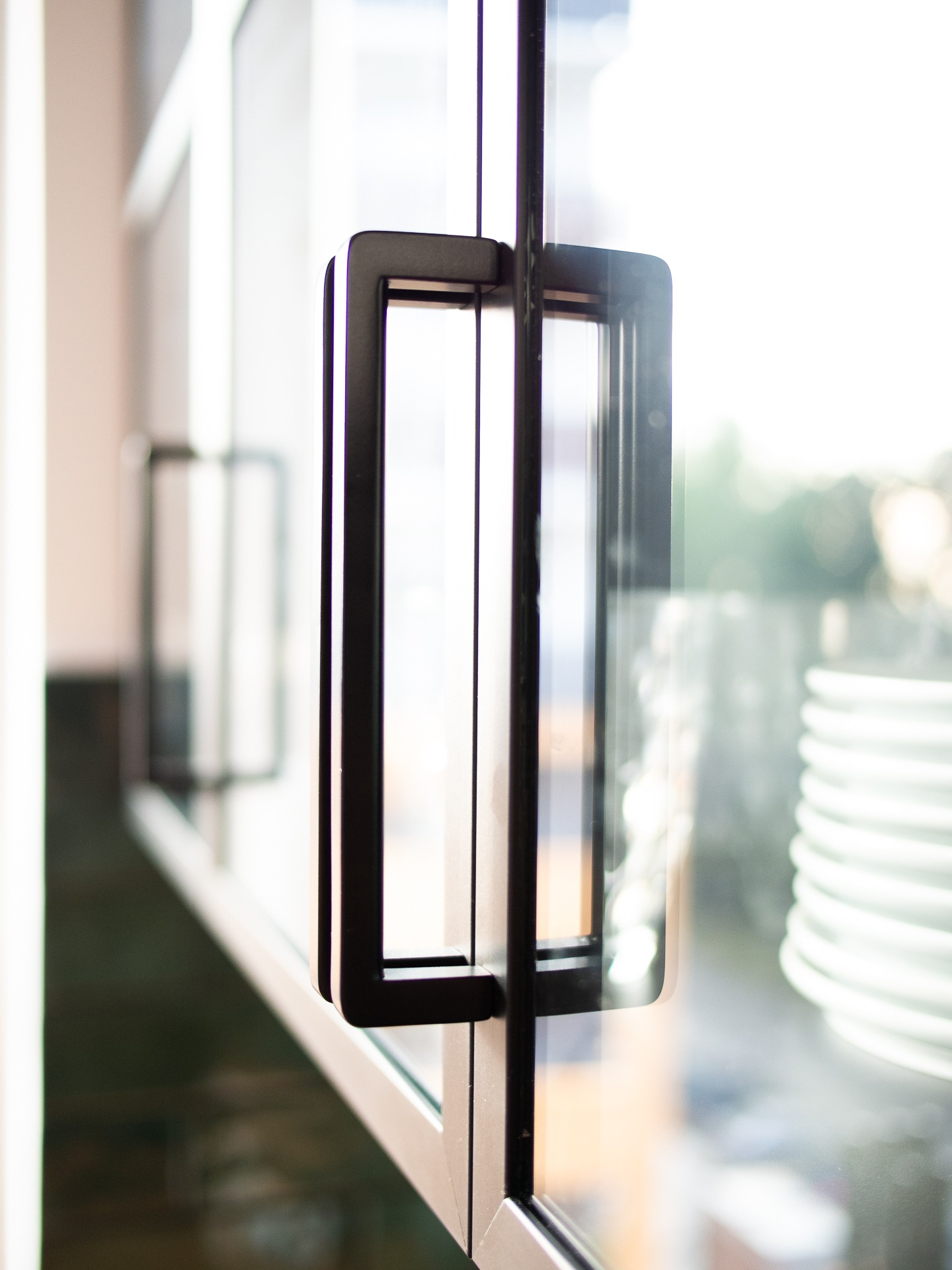 Szklane drzwi z czarnym, prostokątnym uchwytem, widoczne odbicie stosu białych talerzy w szybie.