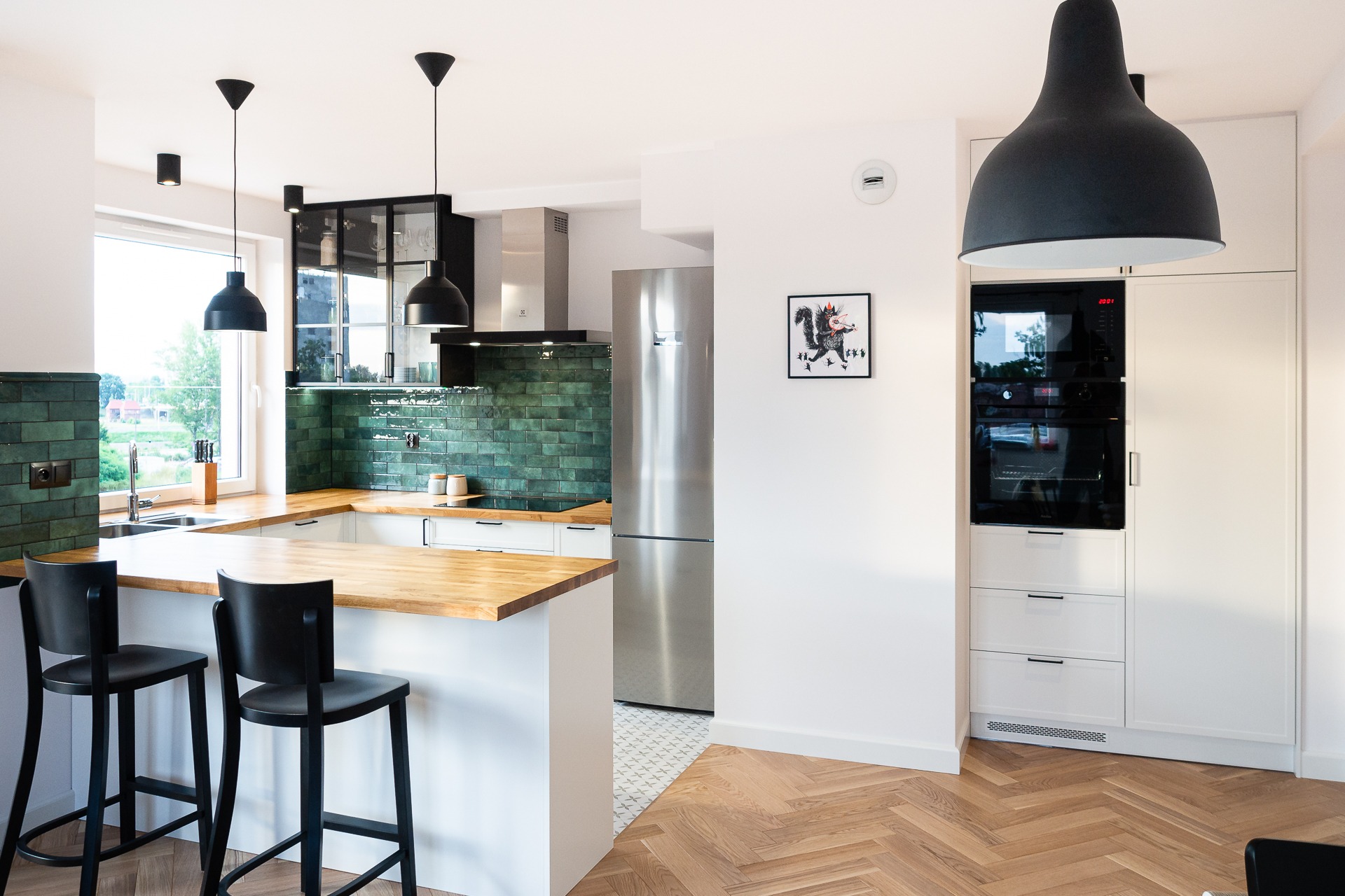 Nowoczesna kuchnia z drewnianym blatem, zielonymi płytkami na ścianie, czarnymi lampami wiszącymi i podłogą ułożoną w jodełkę.