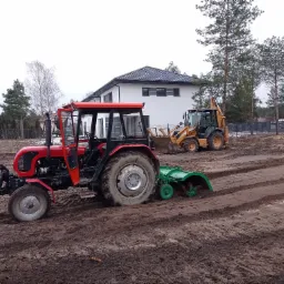 Czerwony traktor z glebogryzarką na polu, w tle żółta koparko-ładowarka i biały dom z czarnym dachem, widoczne ślady pracy w ziemi.