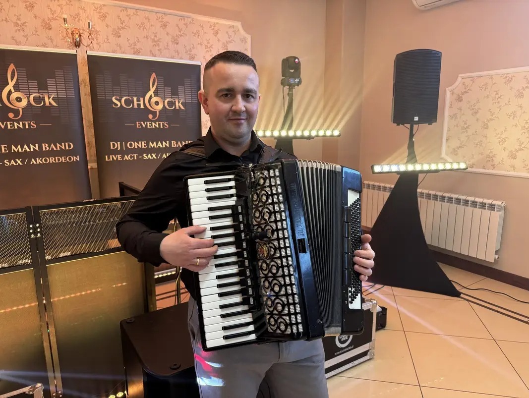 Akordeonista w Mysiadle prezentuje się z instrumentem na tle sprzętu nagłośnieniowego i banerów reklamowych, gotowy do występu na evencie.