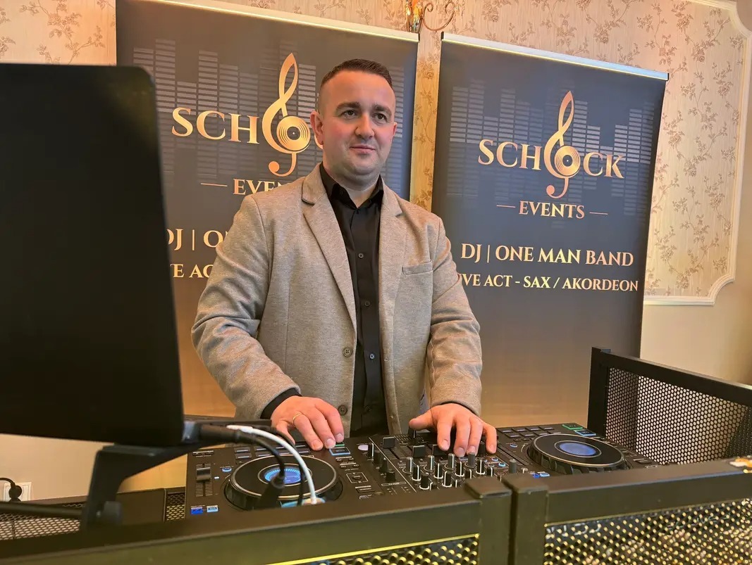 DJ na stanowisku, za nim baner reklamowy Schock Events, oferujący DJ, One Man Band, live act (saksofon, akordeon). Mężczyzna w marynarce, kontroler DJski na pierwszym planie.
