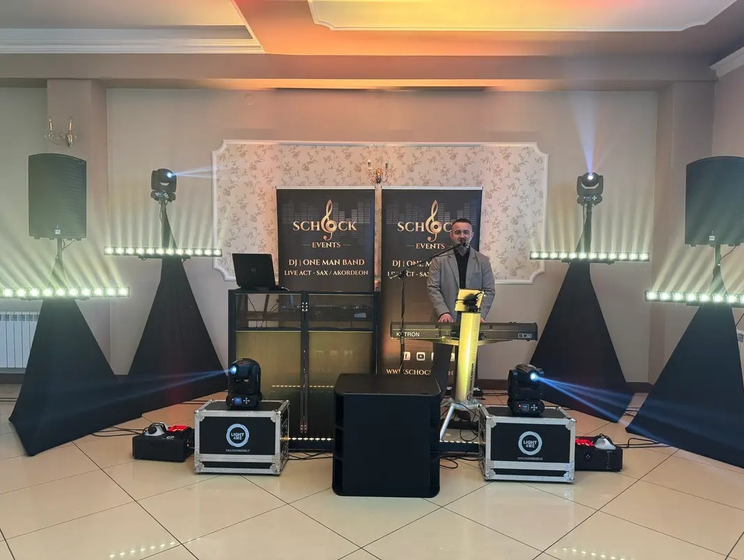 Ustawienie sceniczne z oświetleniem i keyboardem; mężczyzna w marynarce stoi za keyboardem, mikrofon, kolumny, efekty świetlne i banery reklamowe.