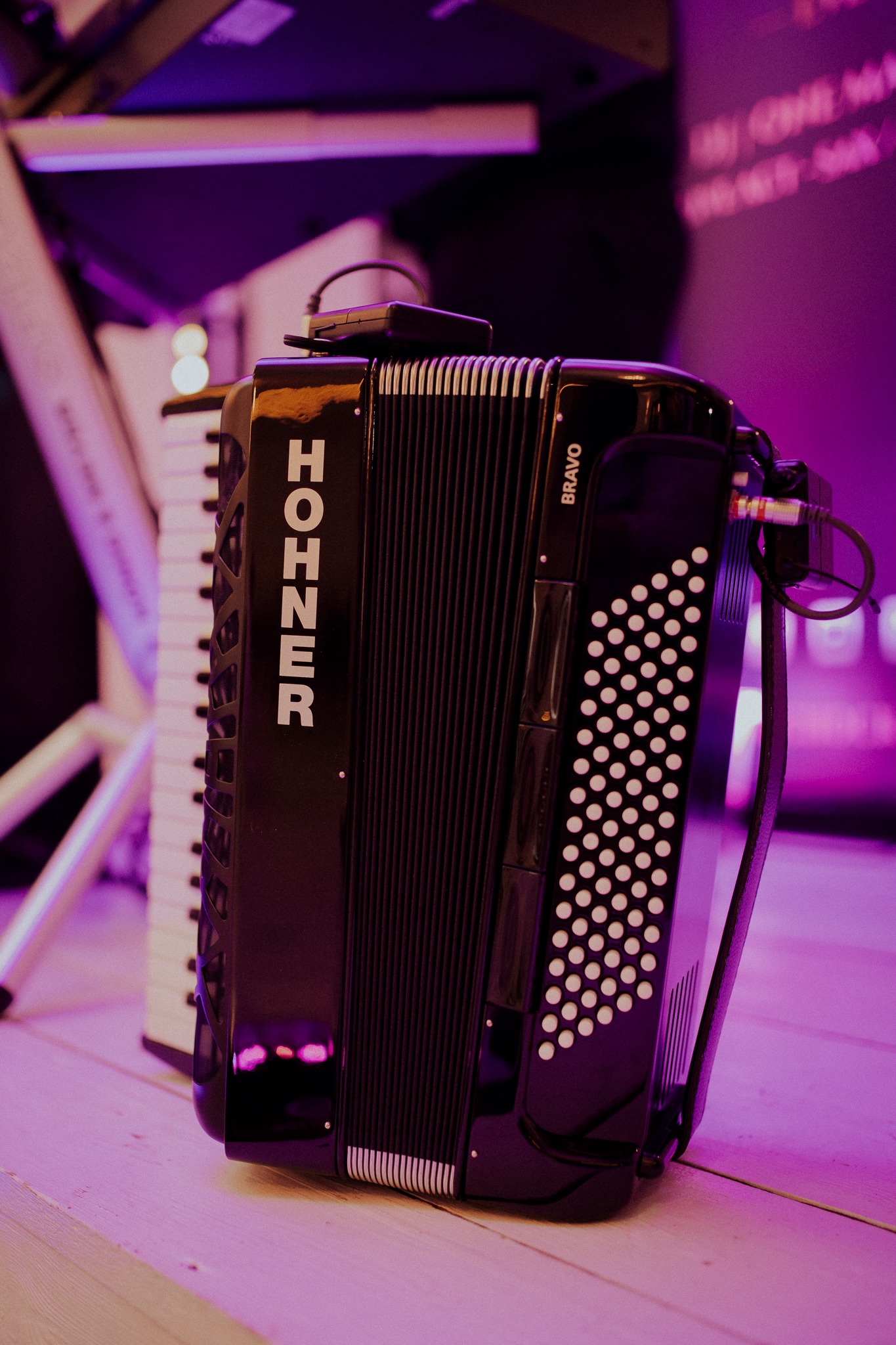 Czarny akordeon Hohner Bravo na drewnianej podłodze, oświetlony fioletowym światłem, z klawiszami w tle. Widoczne kable i elementy instrumentu.