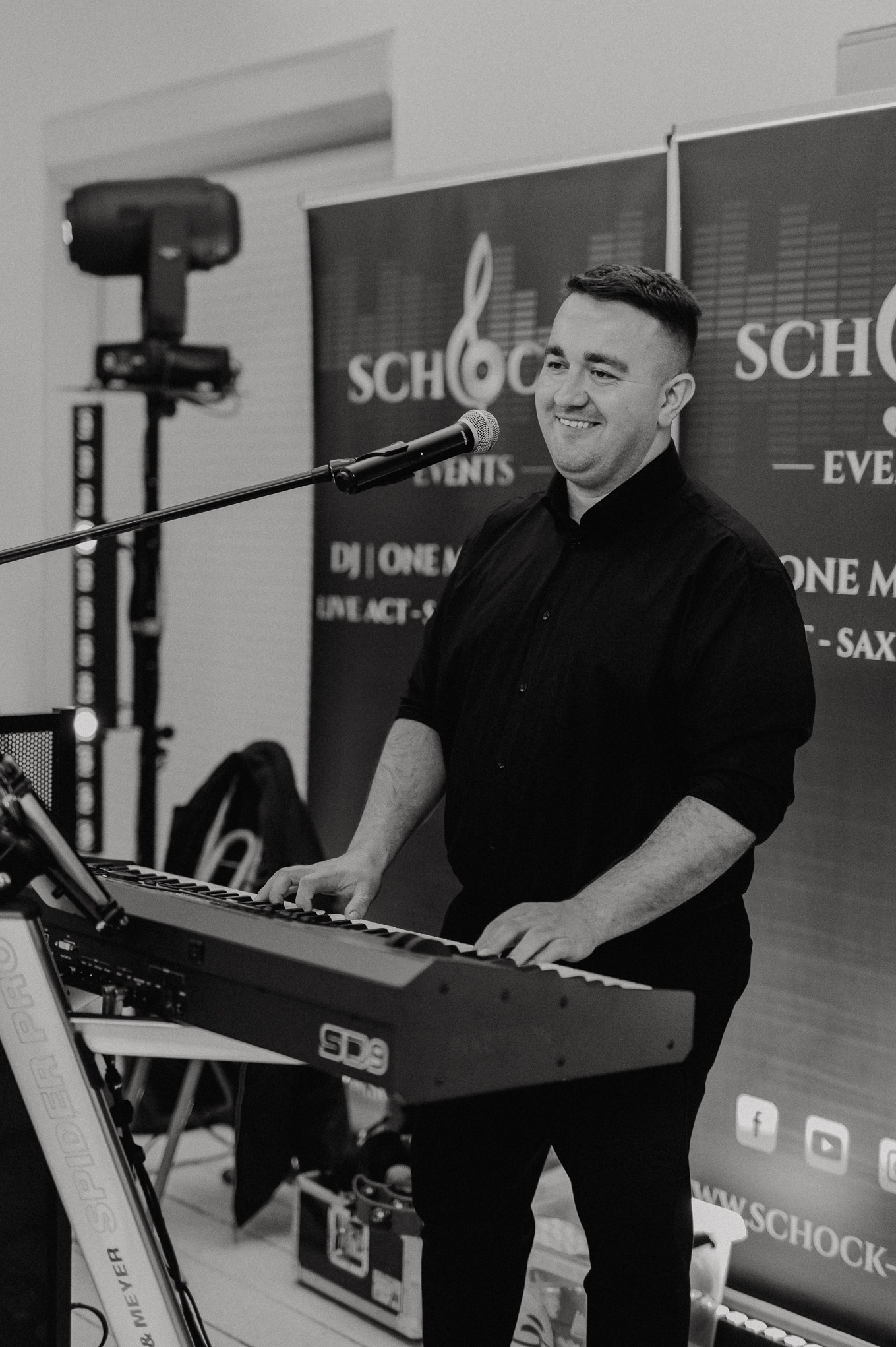 Uśmiechnięty muzyk gra na keyboardzie SD9 podczas występu na tle baneru z logo firmy eventowej i oświetlenia scenicznego. Czarno-biała fotografia.