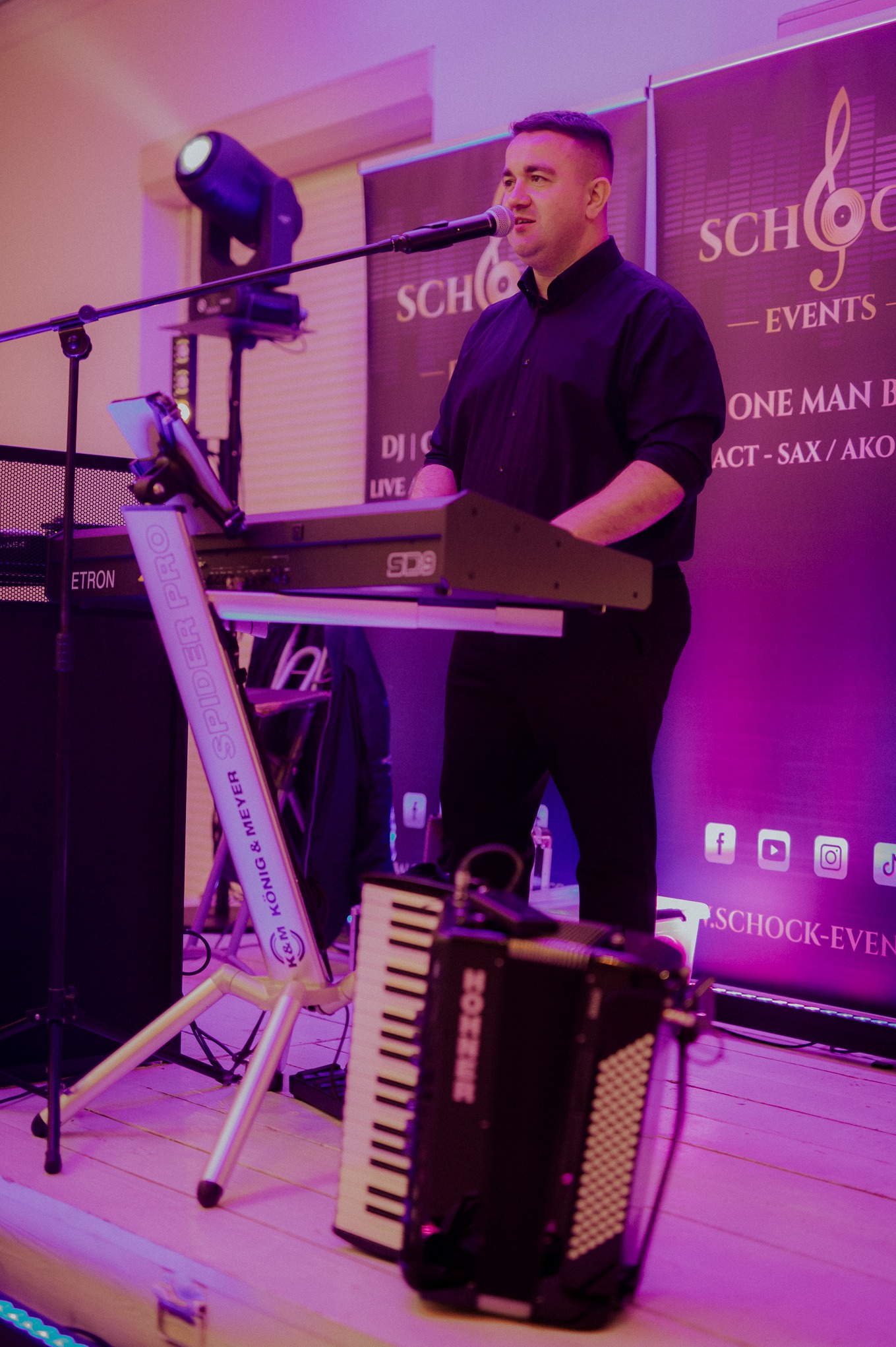 Mężczyzna gra na keyboardzie SD9 na scenie z akordeonem Hohner, oświetlony fioletowym światłem, z banerem Schock-Events w tle. Spider Pro K&M.