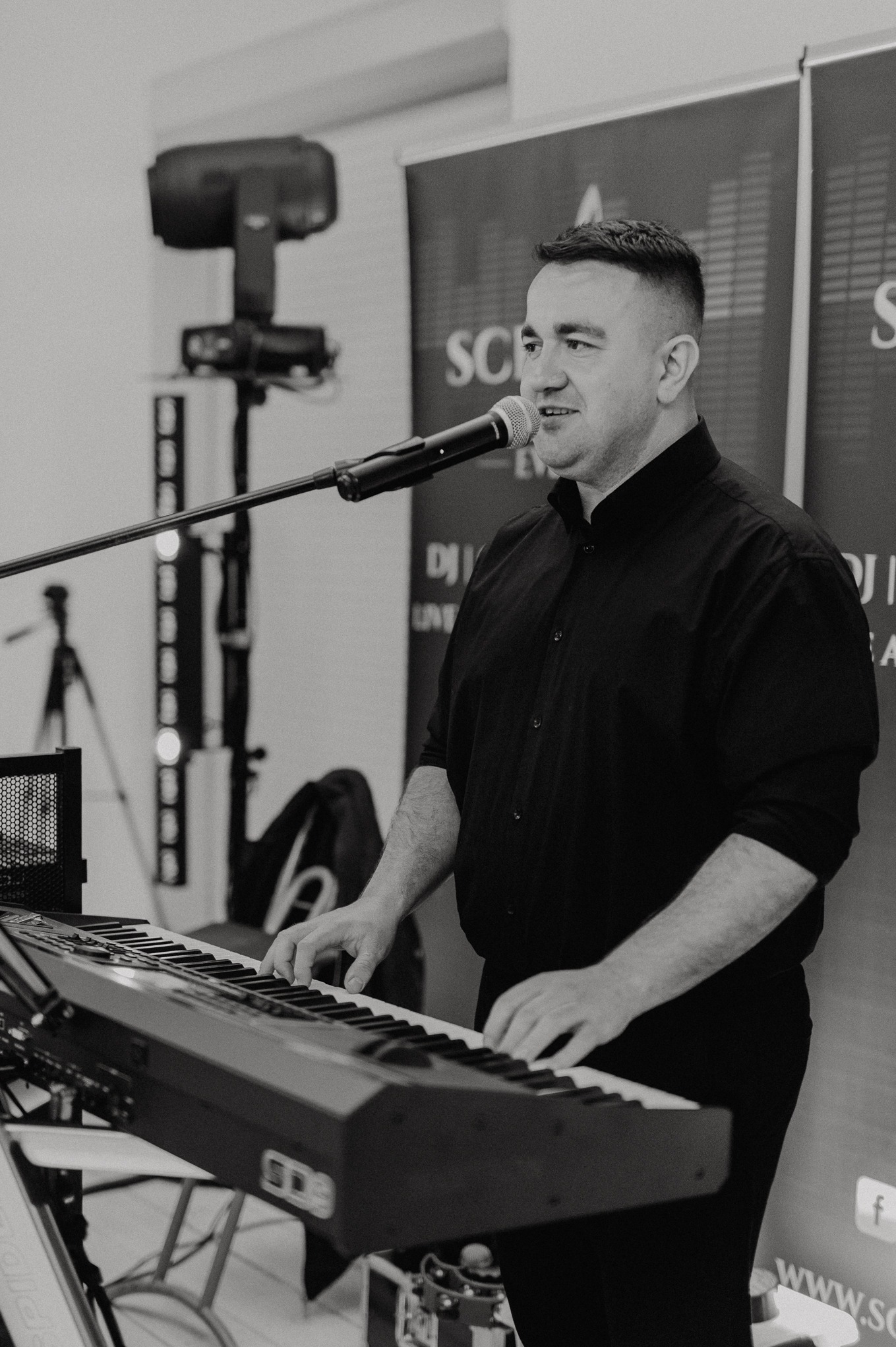 Czarno-białe zdjęcie muzyka grającego na keyboardzie podczas występu, z mikrofonem i oświetleniem scenicznym w tle. Widoczny fragment baneru reklamowego.