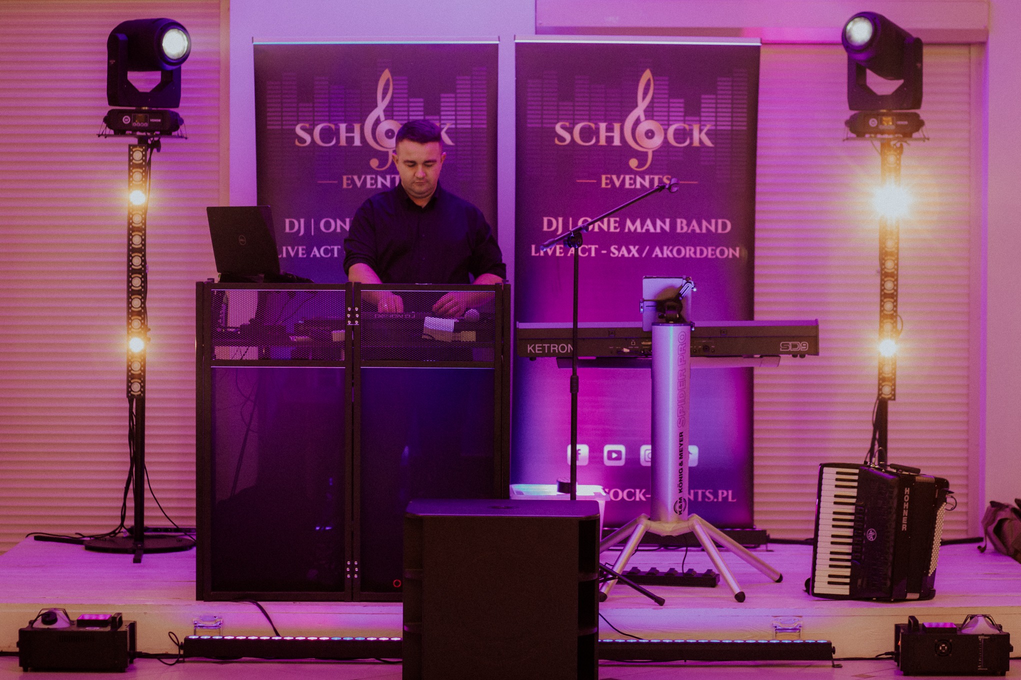 DJ na stanowisku z laptopem i mikserem, keyboard Ketron SD9, akordeon Hohner, oświetlenie sceniczne i banery reklamowe w fioletowym oświetleniu.