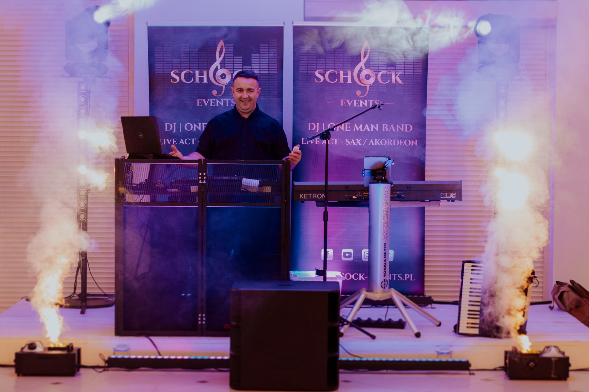 Uśmiechnięty DJ na scenie z laptopem, mikserem, keyboardem Ketron SD9, efektami świetlnymi i dymem. W tle banery z logo 'Schock Events'.