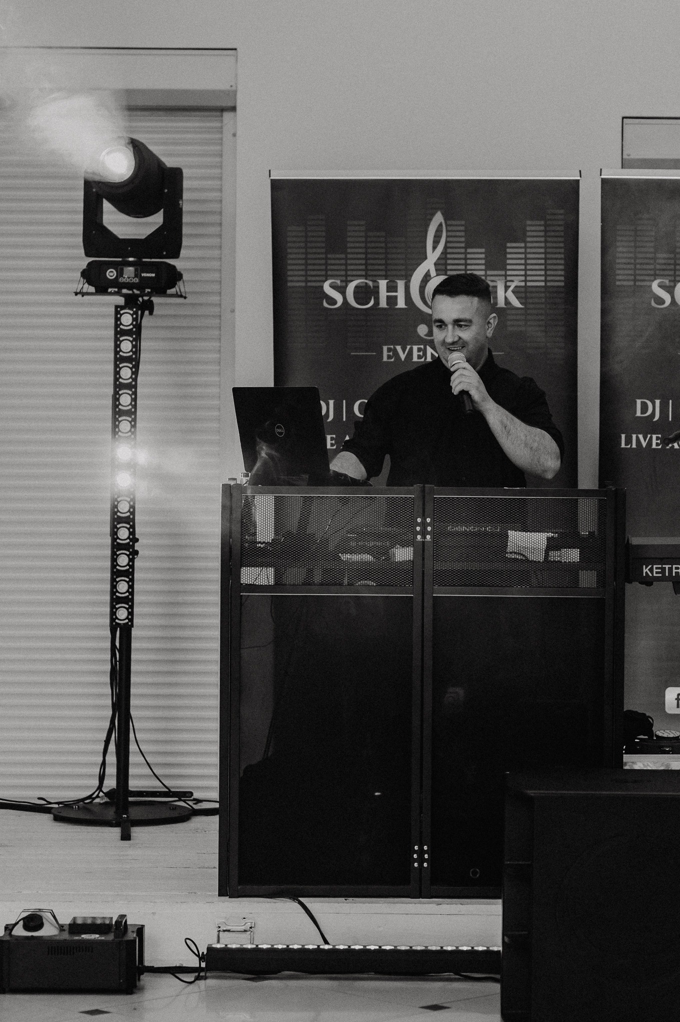 DJ w trakcie występu, oświetlenie sceniczne, laptop Dell, mikrofon, logo firmy na banerze w tle. Czarno-biała fotografia z subtelnym oświetleniem.