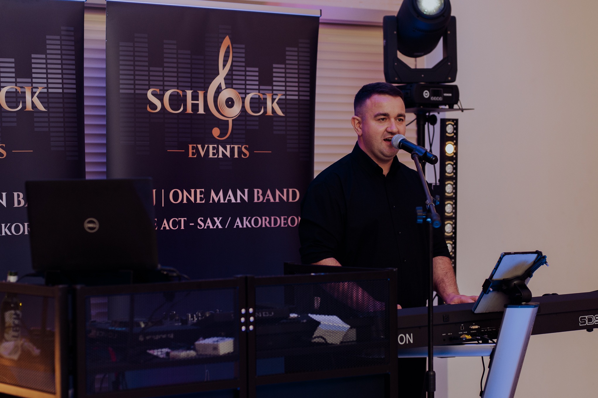 Wokalista z mikrofonem i keyboardem na scenie, za nim baner z logo 'Schock Events', laptop i oświetlenie. Koncertowy występ 'One Man Band'.