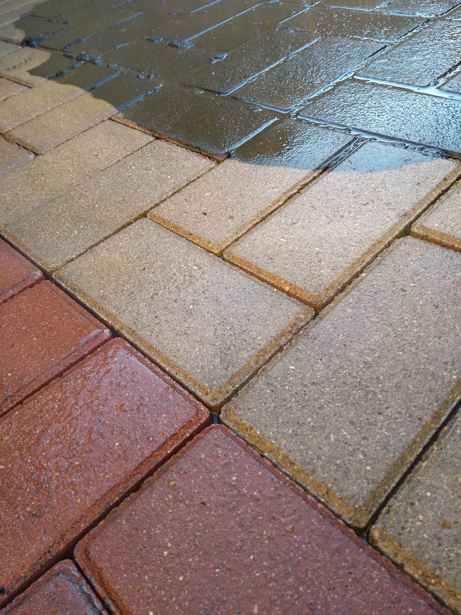 Mokre kostki brukowe w odcieniach czerwieni i szarości, częściowo umyte, z widoczną różnicą w kolorze i czystości między obszarami.