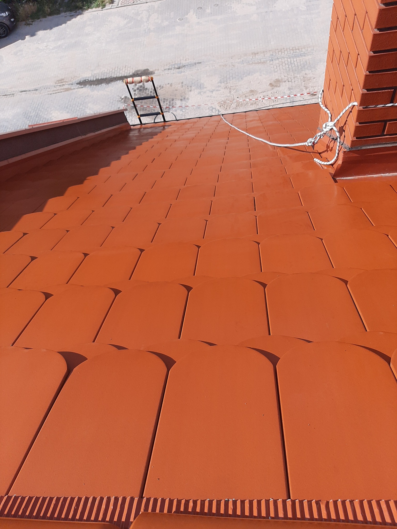 Czerwona dachówka ceramiczna po renowacji, widoczne zabezpieczenie liną przy kominie i drabina w tle.